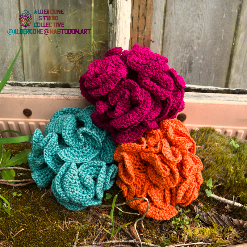 A photo of a stack of three bath poufs on green grass and moss. One of the poufs is maroon-colored, another bright orange, and the last cyan.